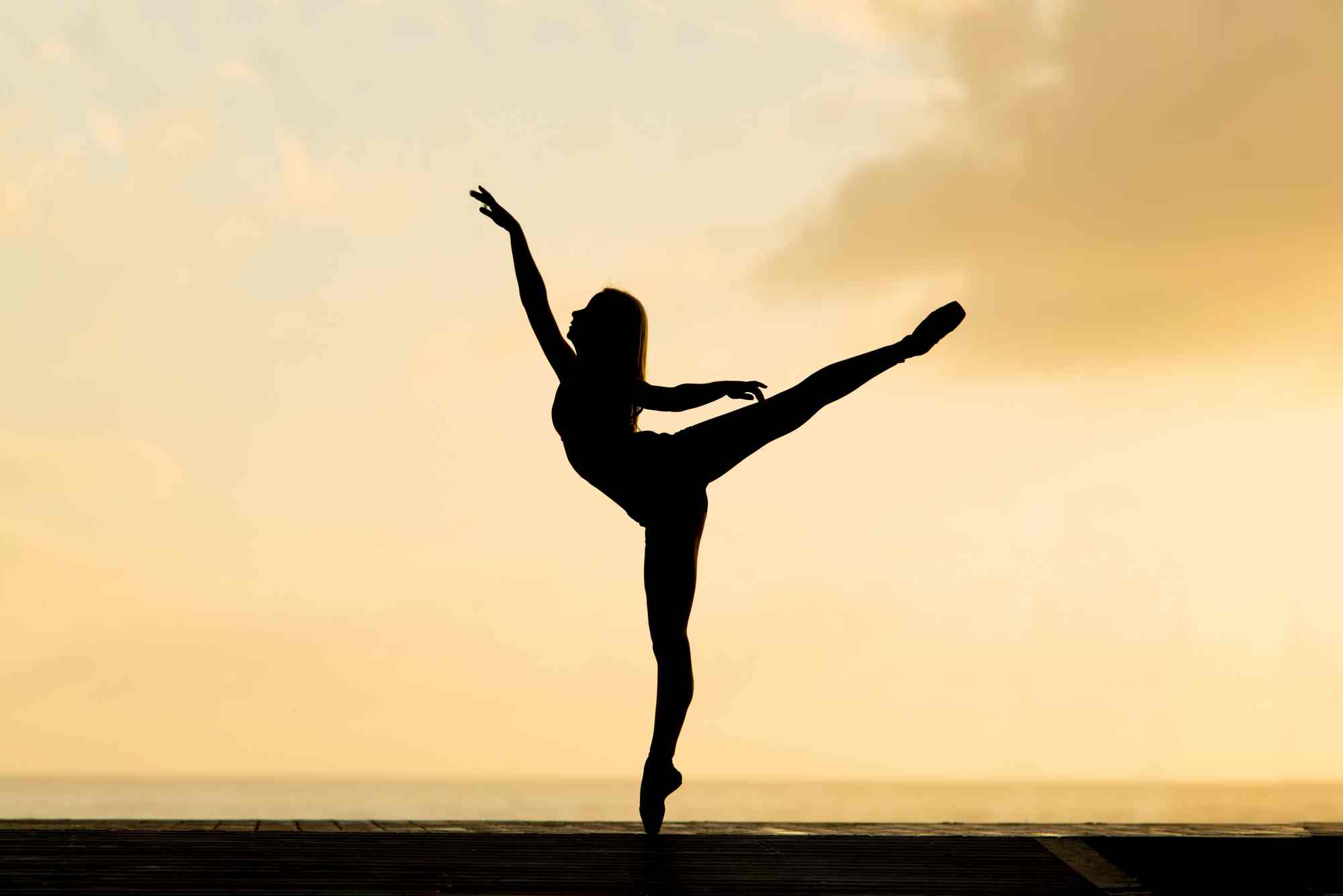 Young lady standing on one leg on tiptoe, as a silhouette before a sunrise sky, arms raised, one leg raised, as if dancing ballet.