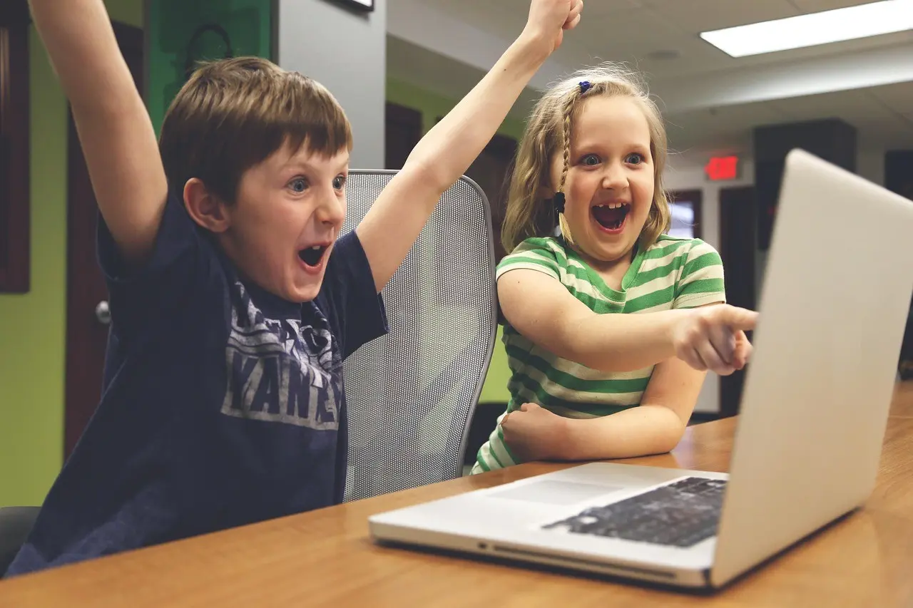 Zwei Kinder vor einem Laptop, die sich sehr freuen, auf den Bildschirm schauend; Bildschirm ist vom Betrachter weggedreht