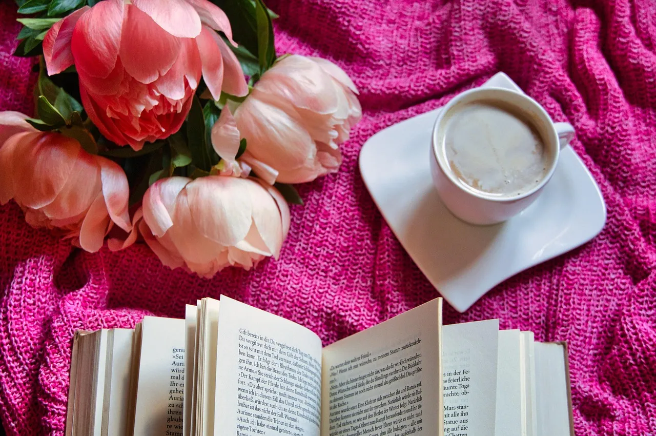 Bild von einem Buch, einer Kaffeetasse und einem Strauß Pfingstrosen auf einem mit pinkfarbener Decke bedeckten Tish.