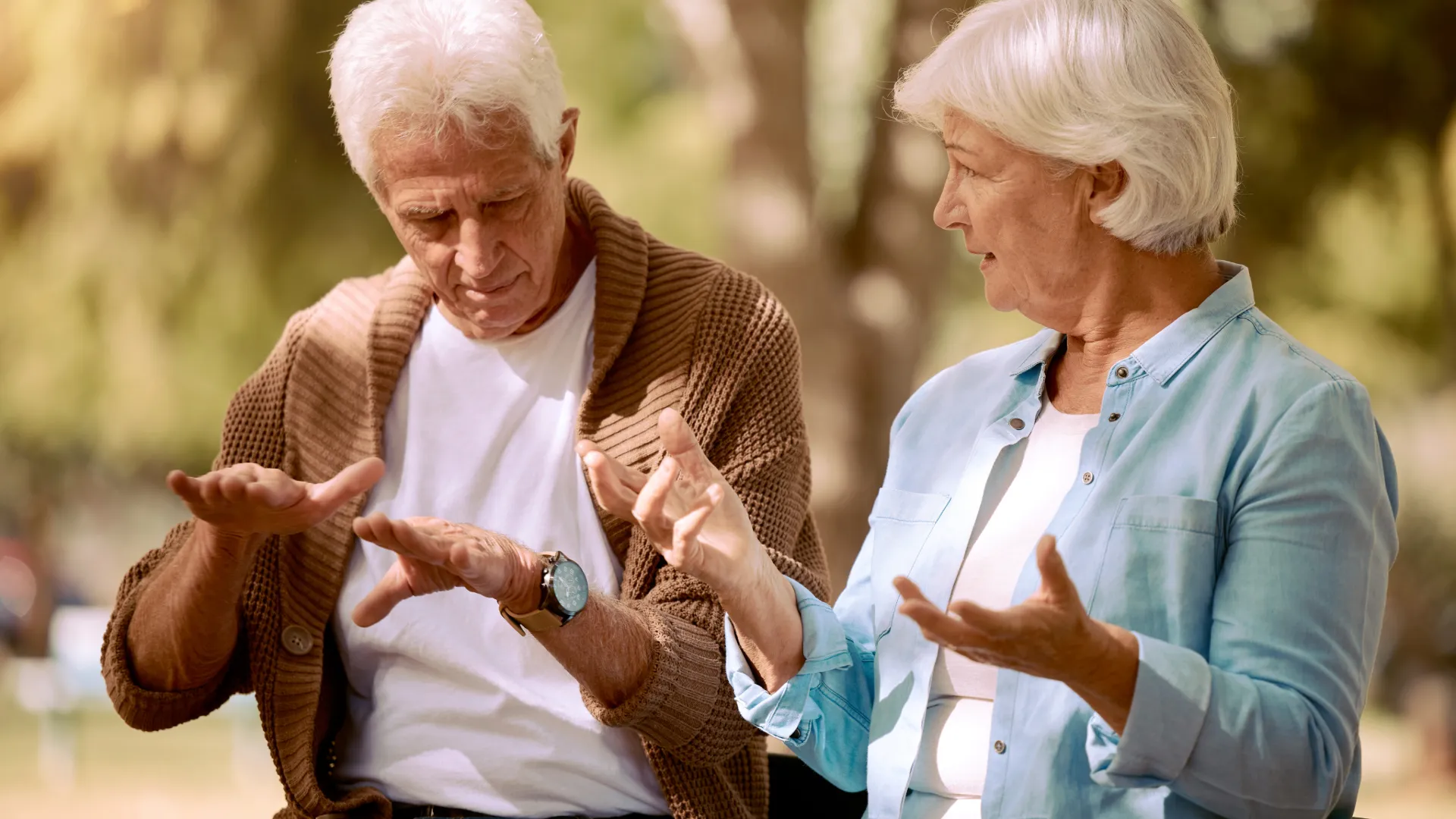 Ein Paar älterer Menschen, die sich auf einer Bank im Park bei sonnigem Wetter sitzend eine freundliche Geste zeigen.
