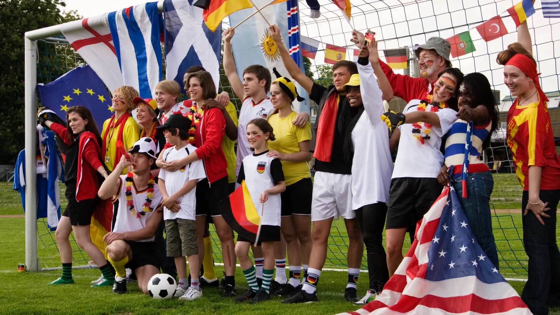 Gruppe von Fußballfans, jung und alt mit verschiedenen Flaggen, die in einem Tor stehen, lächelnd.