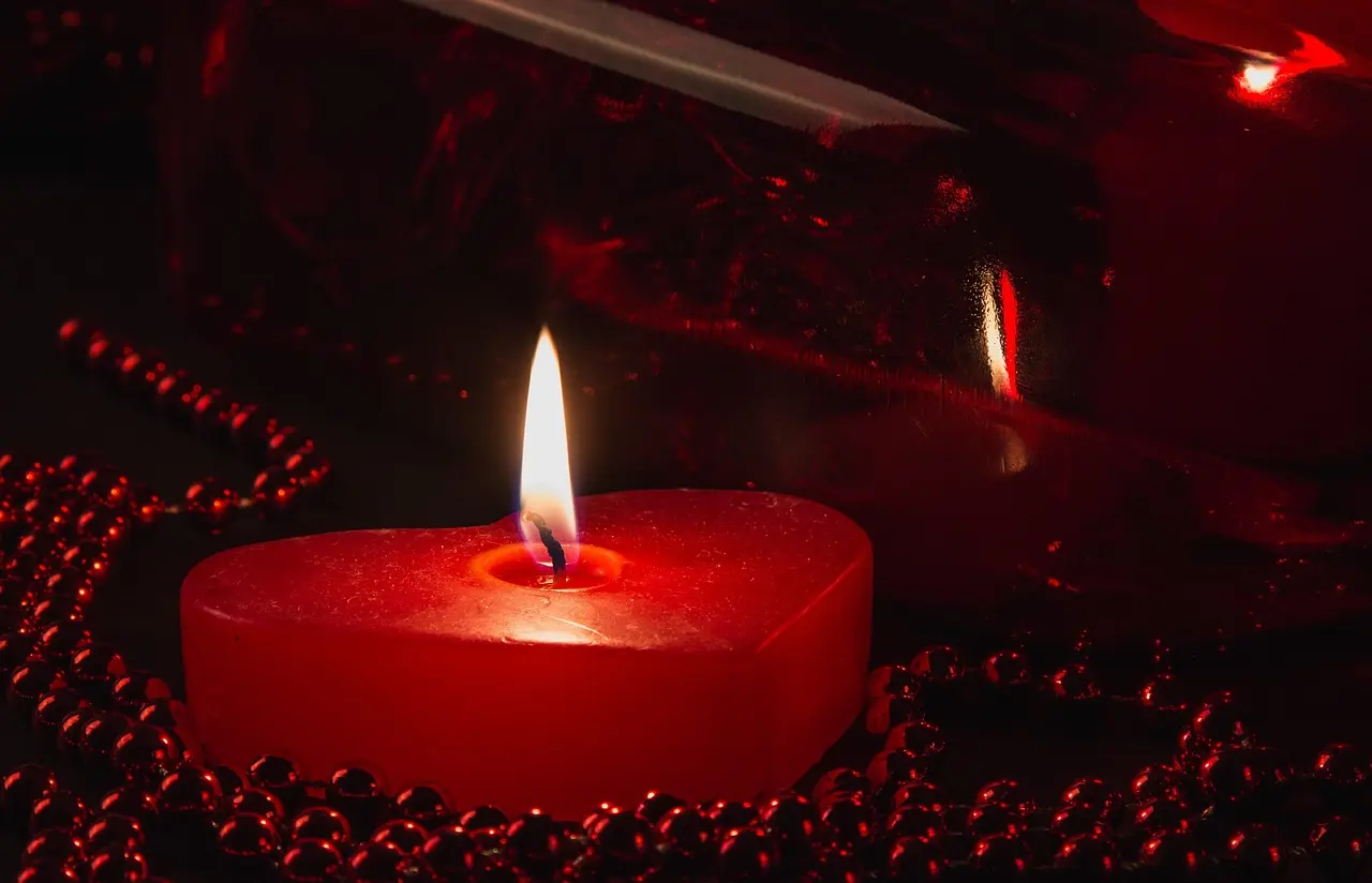 Image of a red, heart-shaped burning candle on a red background, surrounded by many small red and shining beady.