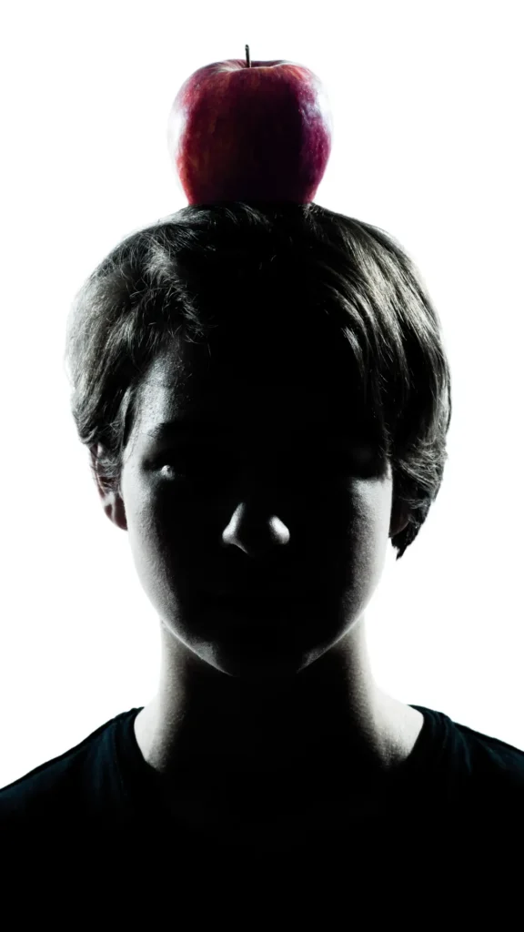 Portraitphoto von jungem Menschen im Schatten, mit Apfel auf dem Kopf.