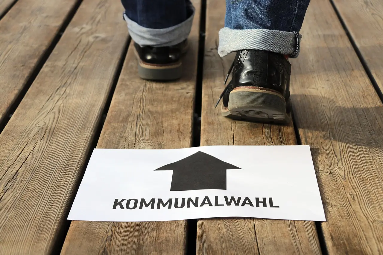 Füße auf einem Holzboden, davor ein Schild, mit Pfeil in Laufrichtung und der Aufschrift: "Kommunalwahl"