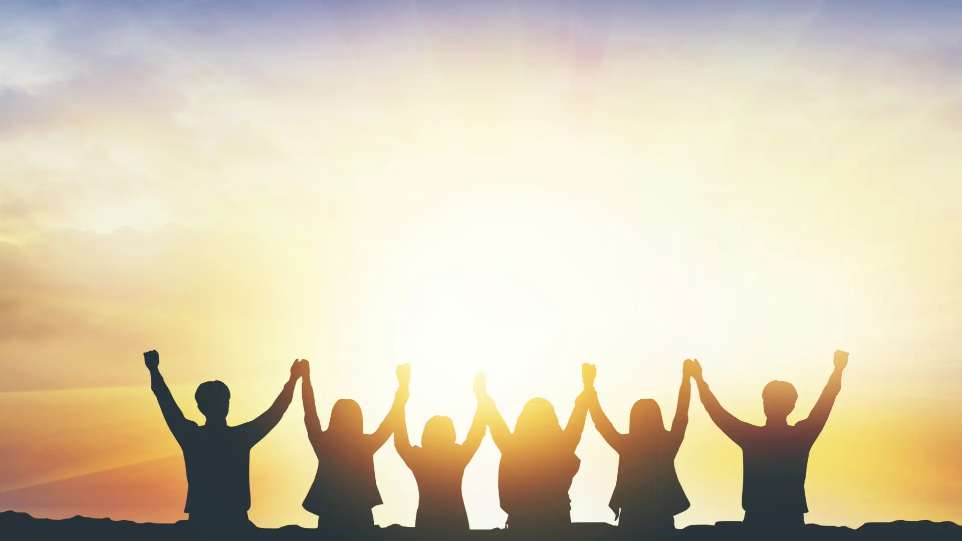 Eine Gruppe von Menschen als Schatten vor einem Sonnenaufgang, die ihre erhobenen Hände aneinanderhalten.